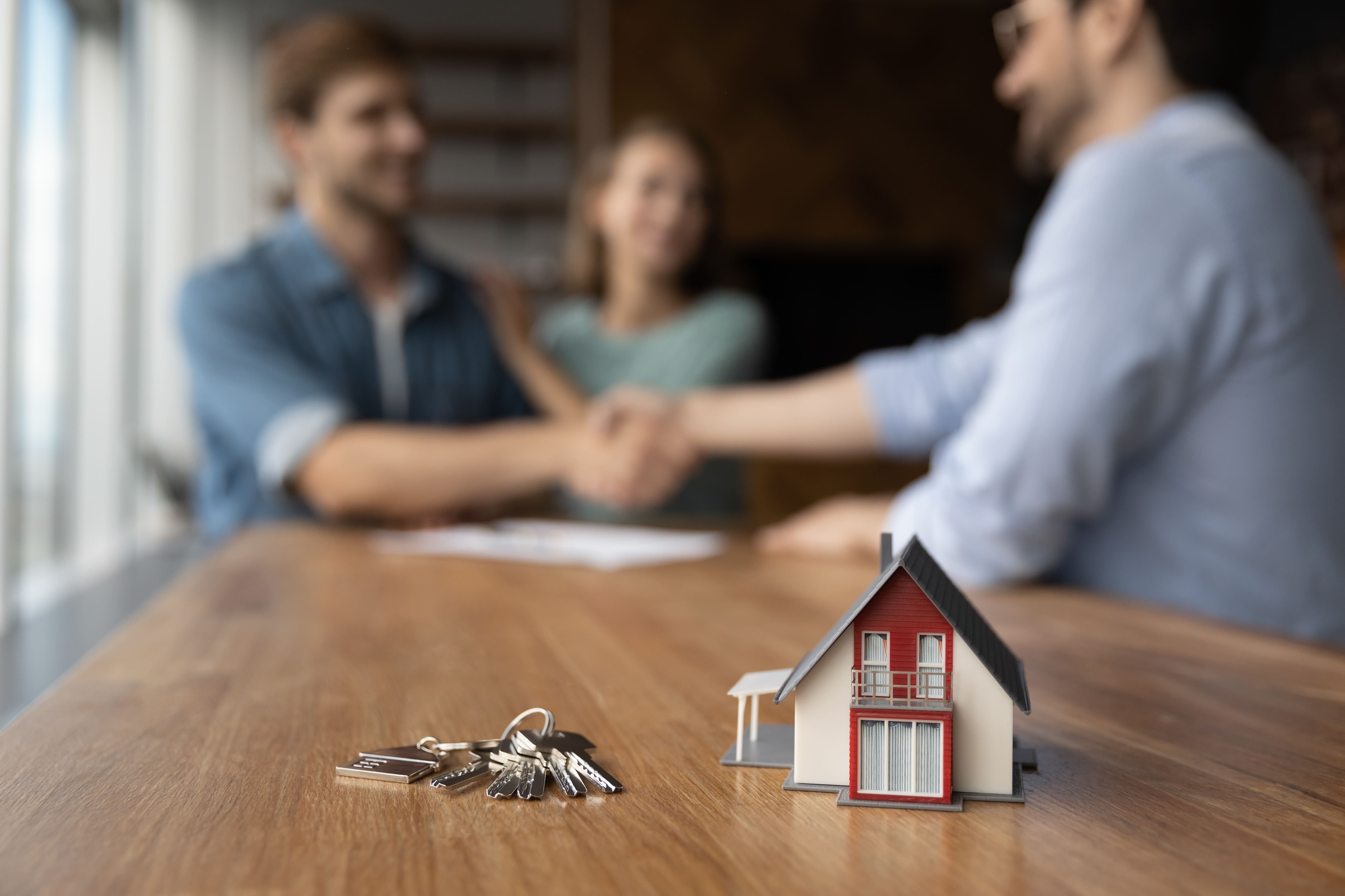 Keys and a small house model on a table with a couple shaking hands with a real estate agent in the background.