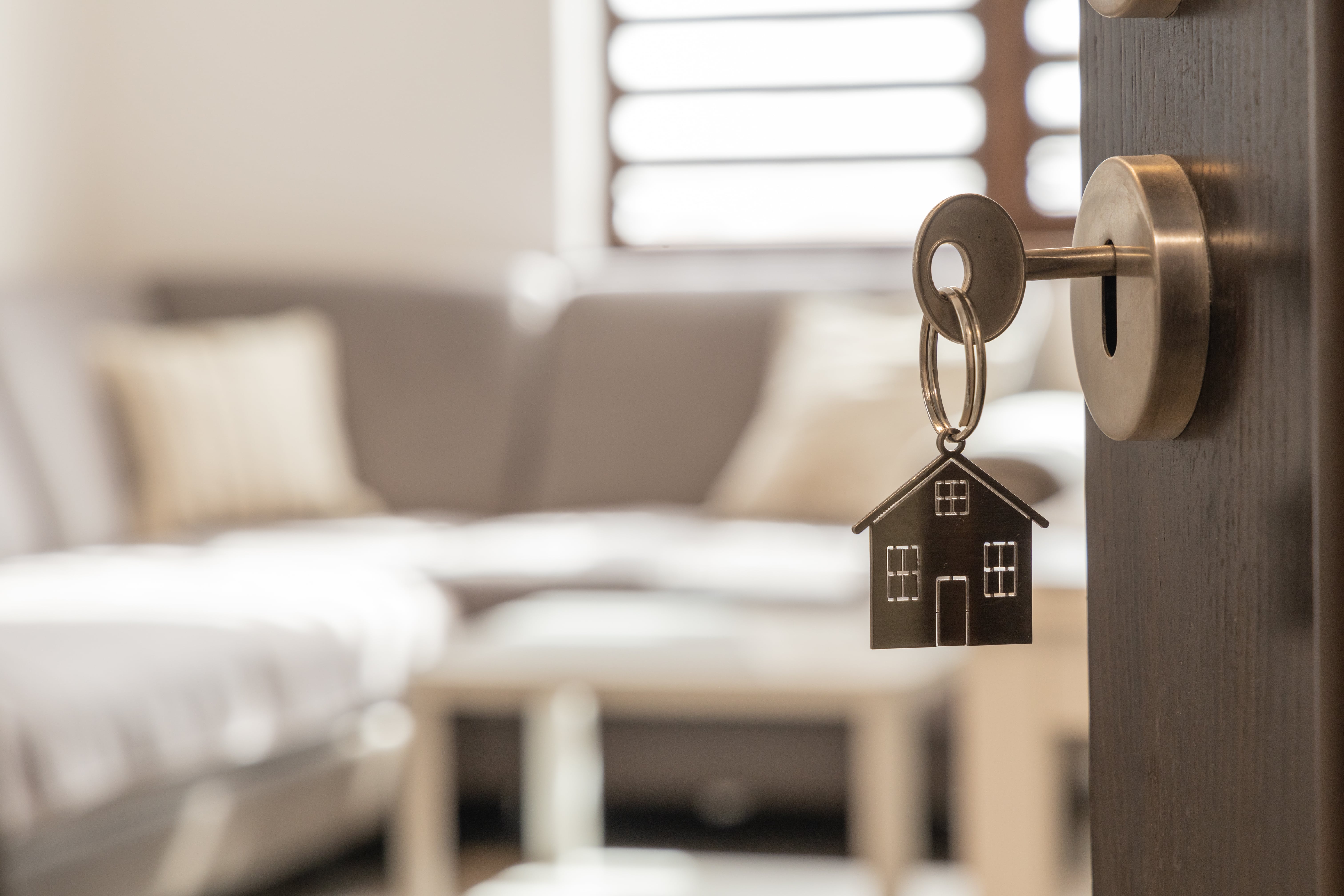 A key with a house-shaped keychain in a door lock, with a living room in the background.